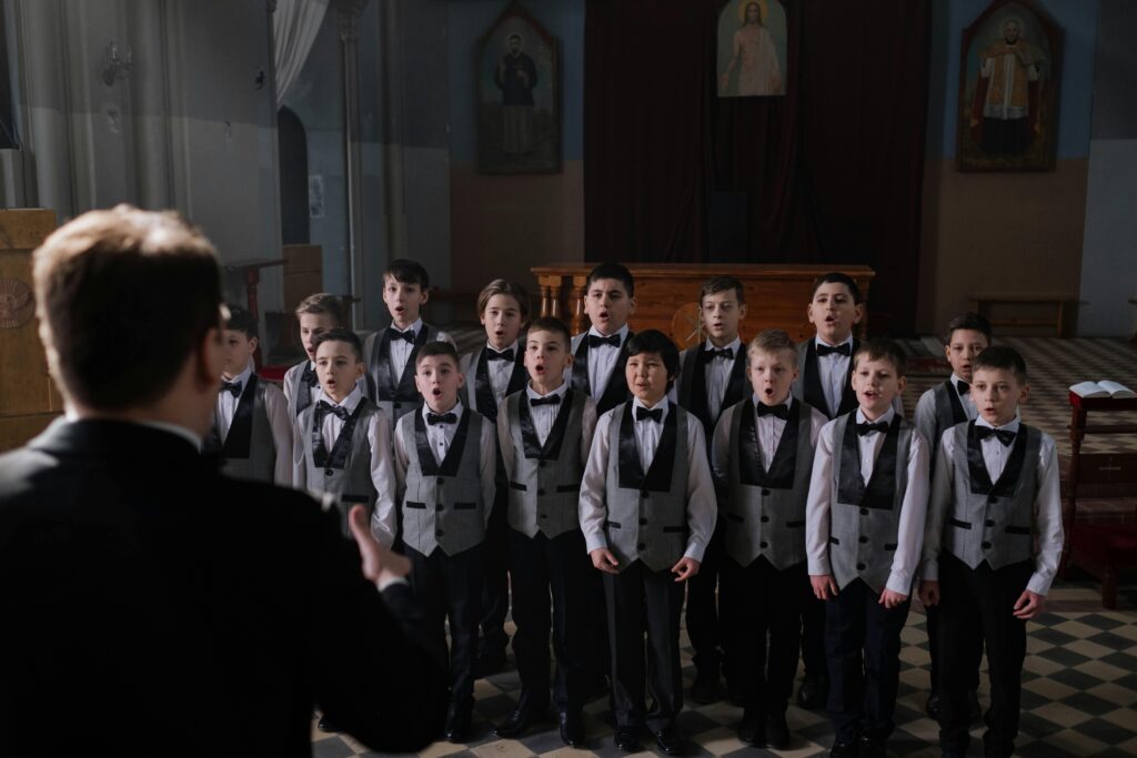 Boys choir practicing indoors, led by conductor, wearing suits.